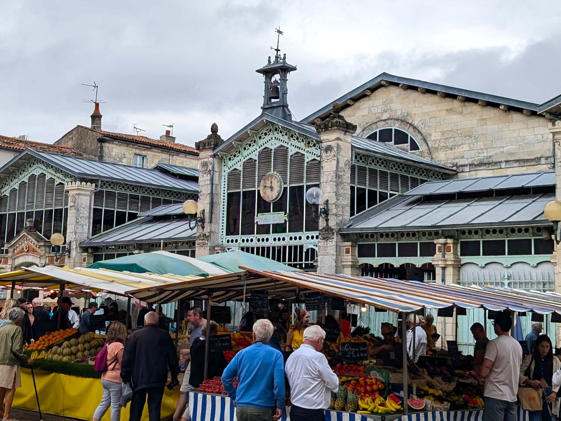 La Rochelle outdoor view of traditional market building, brick and ornate metal frames around glass windows. In front of the building are colourful marketing canopies and people browsing stalls