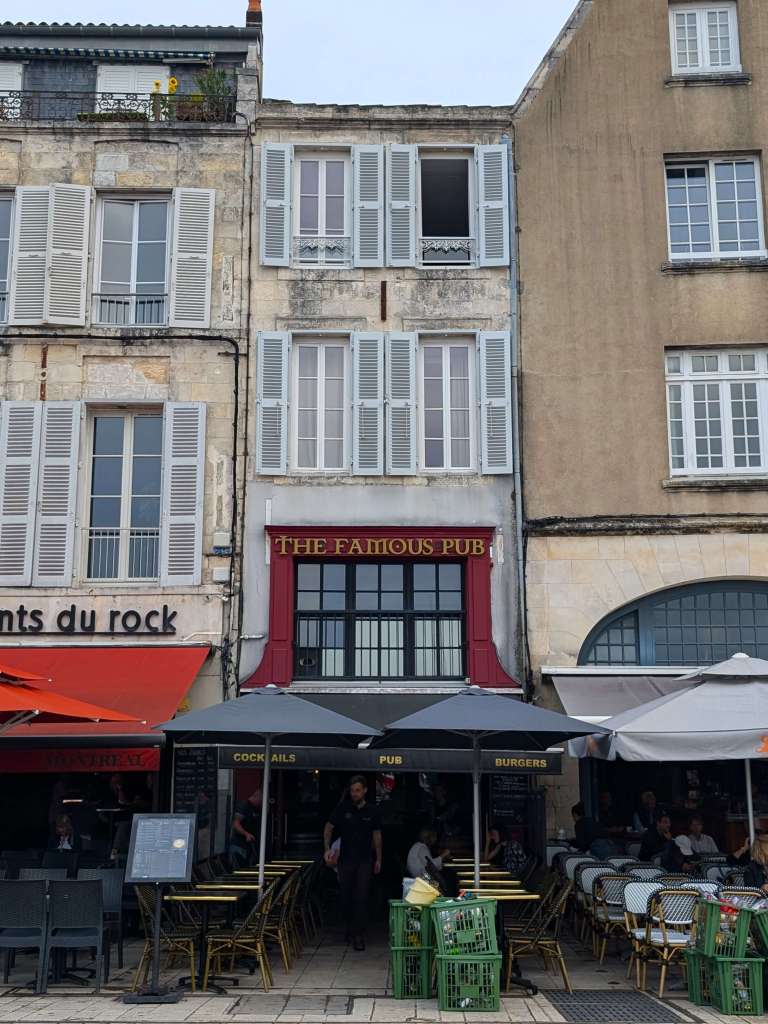 Exterior of The Famous Pub, building is terraced between 2 restaurants. Slatted shutters on the windows above. The Famous Pub sign is gold lettering over red paint.