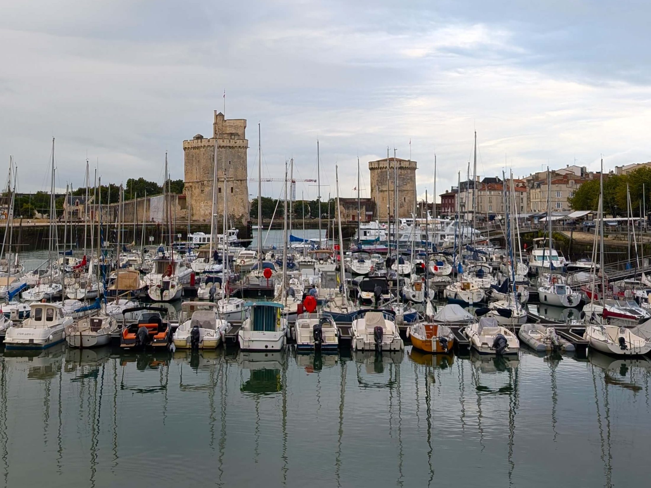 Marina in La Rochelle. Bottom third, dark grey-blue water, middle third approximately 30 small yachts, top third two brick towers and blue sky