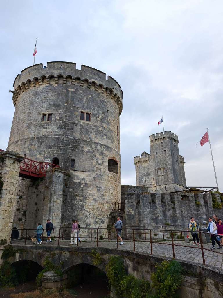 Entrance bridge into archway next to imposing round tower, square tower to the right with a French flag flying. All stone and grey coloured. Blue cloudy sky
